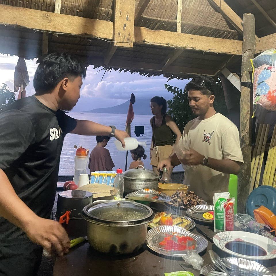 Local Filipino men cooking grilled food under a bamboo shelter by the sea, with travelers and children in the background at sunset on a remote island.