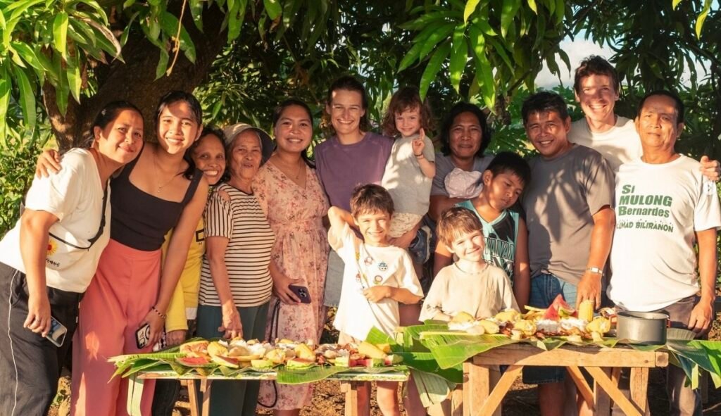 Smiling Filipino family and European travelers sharing a traditional meal under a mango tree during sunset