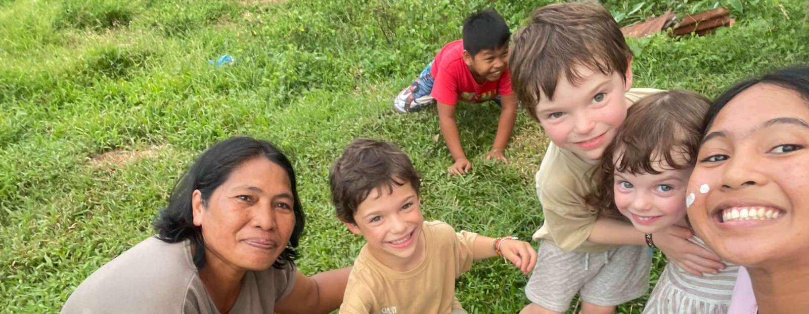 Filipino host and travelers’ children smiling and playing on a green field, gathered around a small black goat, sharing a spontaneous and joyful moment of connection.